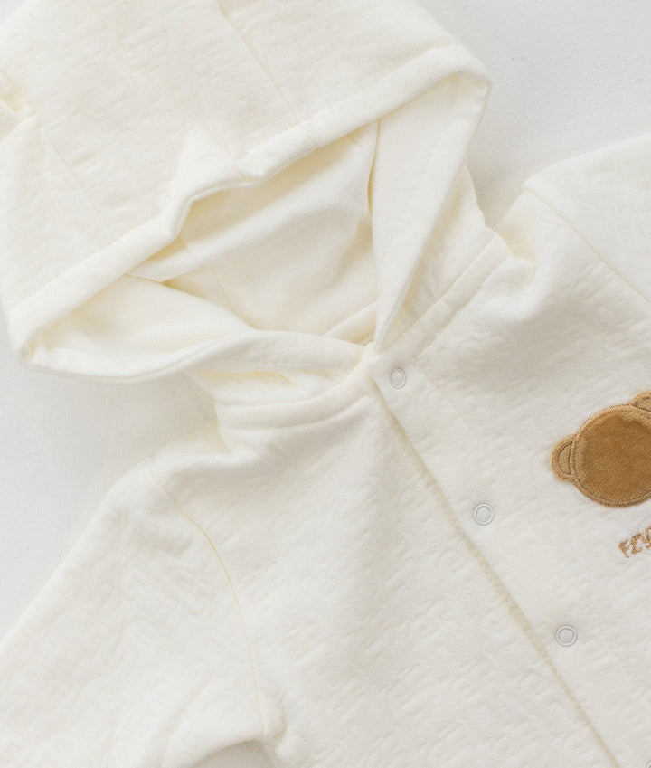 Beige and white hooded bear baby jumpsuit detail showing soft cotton texture, silver snap buttons, and tan embroidered bear face patch