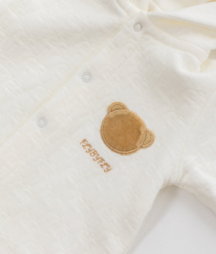 Detail of tan velvet bear head patch and embroidered FLYBYFLY text on left chest of white textured cotton hooded romper