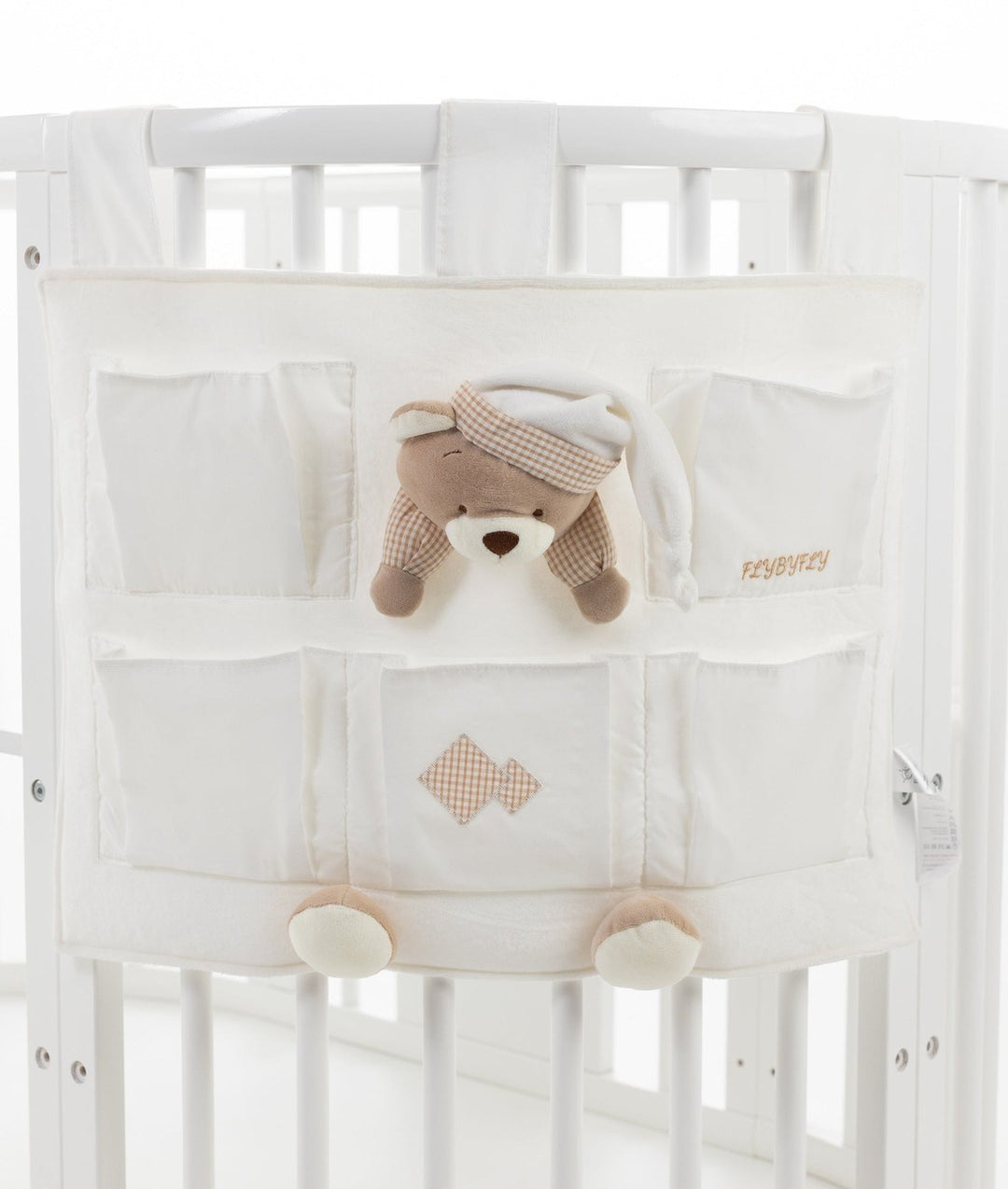Front view of white fabric crib organizer hanging on cot rails with five pockets, beige gingham details, and central plush bear